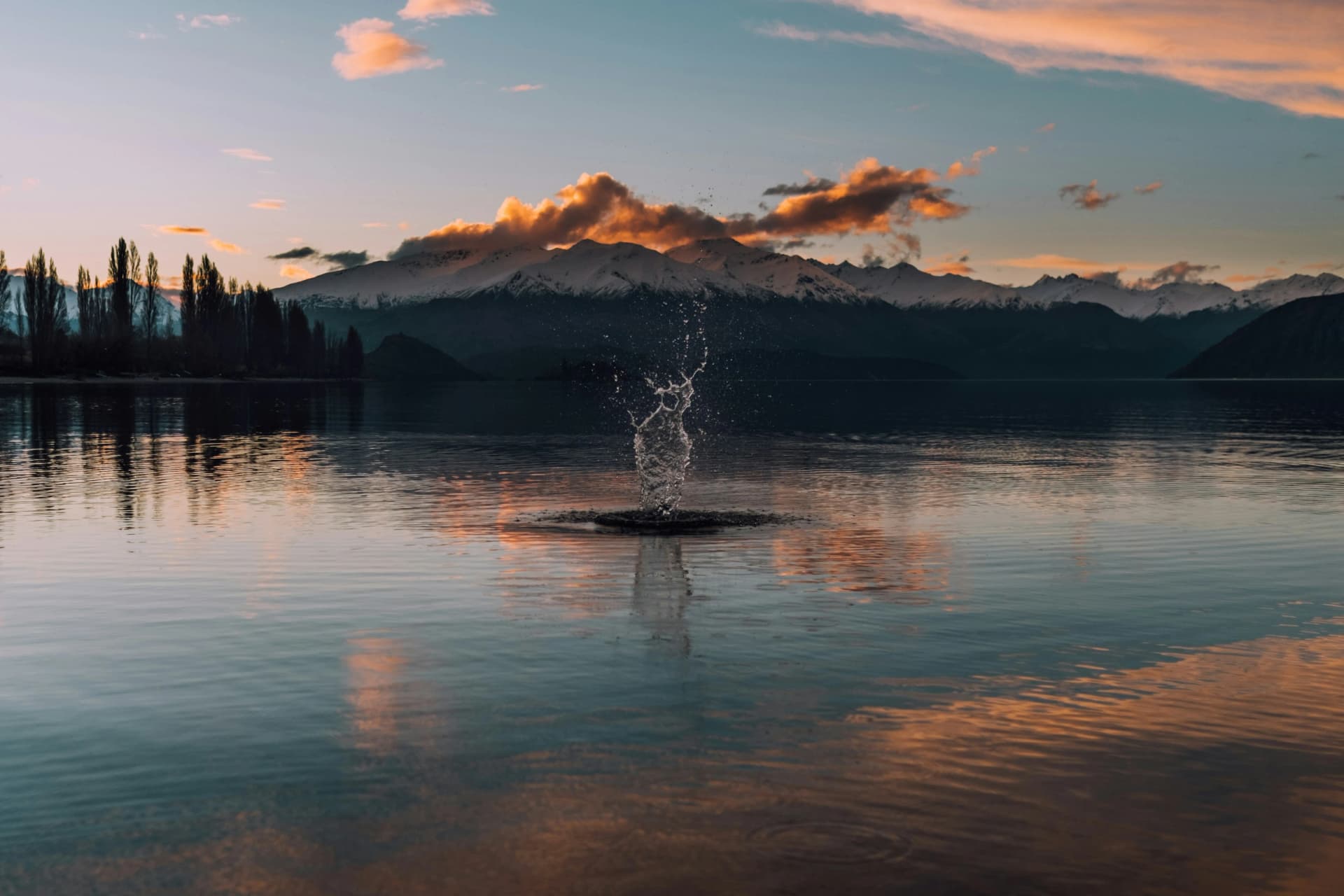 Water splashing up from the calm surface of a lake at sunset, with snow-capped mountains and pink clouds reflected behind it